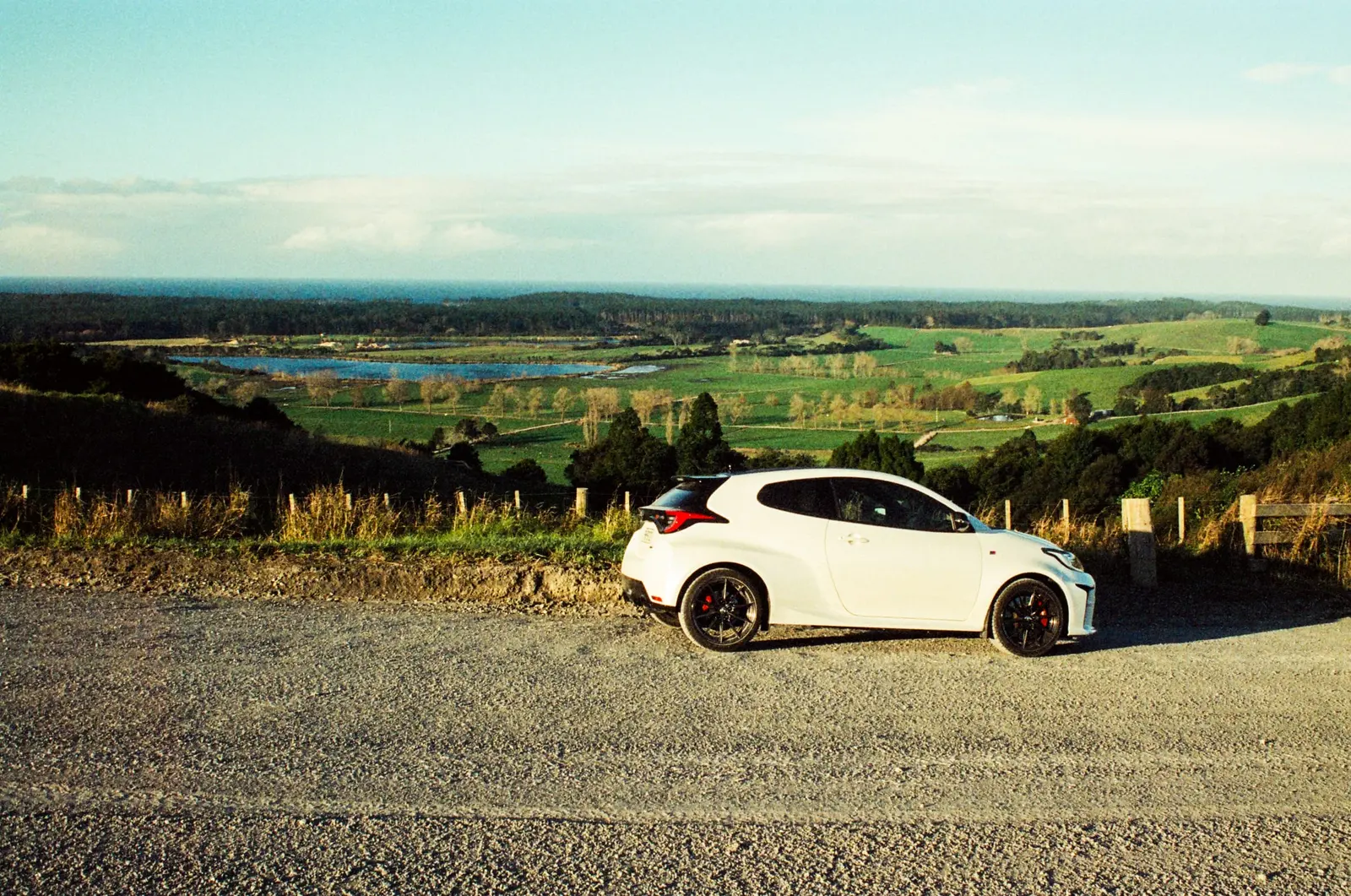 White Toyota GR Yaris on a gravel lookout above green farmland and a lake, in late-afternoon light